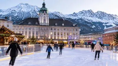 KI-Symbolbild: Menschen laufen Schlittschuh auf einer beleuchteten Eisfläche vor einem historischen Gebäude und verschneiten Bergen.