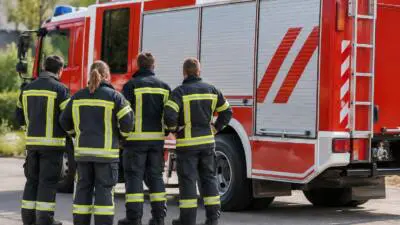 Personen in dunkler Schutzkleidung mit gelben Reflexstreifen stehen neben einem roten Einsatzfahrzeug.