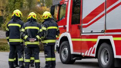 KI-generiertes Bild: Feuerwehrleute in dunkler Schutzkleidung stehen neben einem roten Feuerwehrfahrzeug.