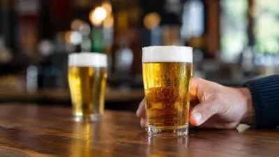 KI-Symbolbild: Eine Hand hält ein schmales Glas mit goldenem Bier auf einer Holztheke.