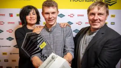 Ein junger Mann präsentiert eine Trophäe vor einer Sponsorenwand bei einer Hochbau-Veranstaltung.
