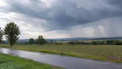 KI-Symbolbild: Dunkle Regenwolken über Feldern, Regen fällt auf eine Landstraße.