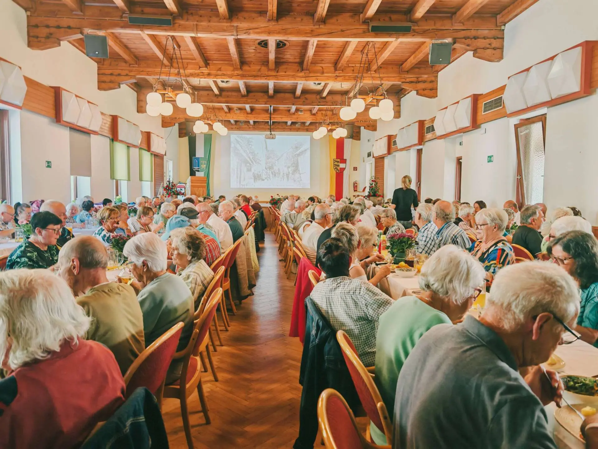 Ältere Gäste sitzen an gedeckten Tischen, während eine historische Aufnahme auf die Leinwand projiziert wird.