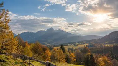 KI-generiertes Bild: Eine herbstliche Berglandschaft mit gelben Bäumen, Wolken und Sonne über einem Tal.