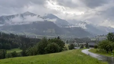 KI-generiertes Bild: Bewölkte Berglandschaft mit tief hängenden Wolken, grüner Wiese und nasser Straße