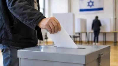 KI-Symbolbild: Eine Hand steckt einen Stimmzettel in eine Wahlurne; im Hintergrund hängt eine israelische Flagge.