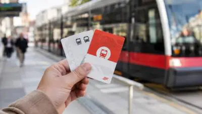 KI-Symbolbild: Eine Hand hält Fahrkarten vor einer Straßenbahn am Bahnsteig.