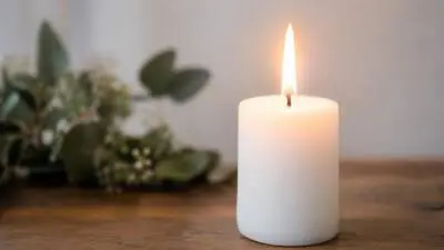 KI-Symbolbild: Eine brennende weiße Kerze steht auf einem Holztisch vor unscharfen Blumen.