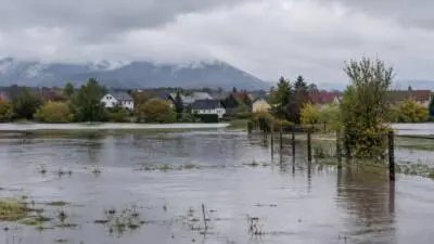 KI-Symbolbild: Überflutete Wiesenfläche vor einem Dorf mit Häusern und bewölkten Bergen.