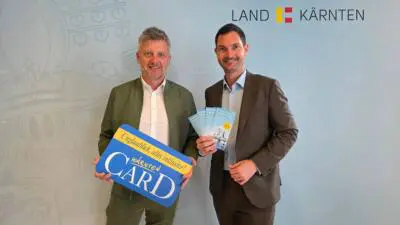 Zwei Männer präsentieren eine Kärnten-Card-Tafel und Folder zum Winterfreizeitpass.