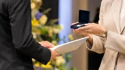 KI-Symbolbild: Hände in formeller Kleidung bei der Übergabe einer Medaille und eines Dokuments.