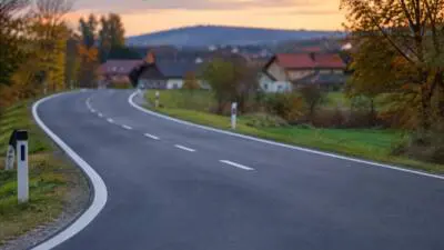 KI-generiertes Bild: Eine asphaltierte Straße mit weißen Markierungen führt in einer Kurve durch eine Ortschaft.