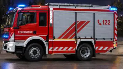 KI-generiertes Bild: Ein rotes Feuerwehrfahrzeug mit Blaulicht steht auf nasser Fahrbahn.