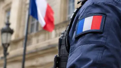 KI-Symbolbild: Eine dunkelblaue Uniform mit französischer Flagge vor unscharfer Fassade und Trikolore.