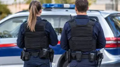 KI-Symbolbild: Zwei Personen in dunkler Uniform und Schutzwesten vor einem weiß-blauen Auto mit Blaulicht.