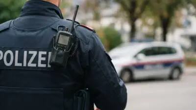 KI-Symbolbild: Ein Polizist mit Funkgerät vor einem Streifenwagen.