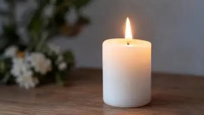KI-generiertes Bild: Eine brennende weiße Stumpenkerze steht auf einer Holzfläche, daneben unscharfe Blumen.
