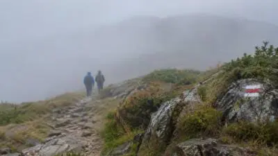 KI-generiertes Bild: Wandernde auf einem felsigen Bergpfad im Nebel neben einer rot-weißen Wegmarkierung.