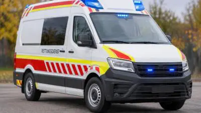 KI-generiertes Bild: Ein weißer Rettungswagen mit rot-gelben Streifen und eingeschaltetem Blaulicht steht auf einer befestigten Fläche.