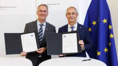 Zwei Männer in Anzügen präsentieren geöffnete Dokumentenmappen neben einer EU-Flagge.