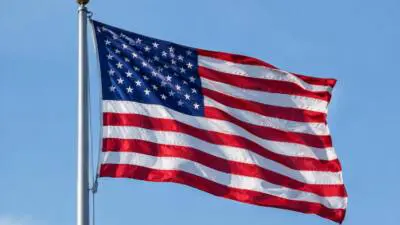 KI-Symbolbild: Eine amerikanische Flagge weht an einem Mast vor blauem Himmel.