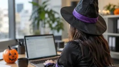KI-Symbolbild: Eine Person mit Hexenhut sitzt vor einem Laptop; ein Halloweenkürbis steht auf dem Schreibtisch.