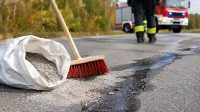 KI-Symbolbild: Streugut und Besen neben einer dunklen Flüssigkeitsspur auf einer Straße.