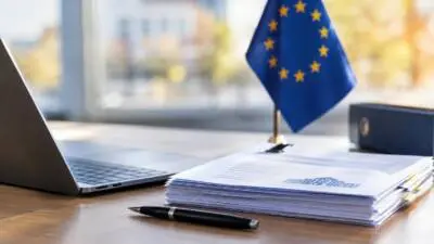 KI-Symbolbild: Eine EU-Flagge neben Laptop und Unterlagen auf einem Schreibtisch.