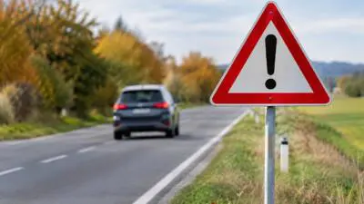 KI-Symbolbild: Ein rotes dreieckiges Warnschild steht neben einer Landstraße, dahinter fährt ein graues Auto.