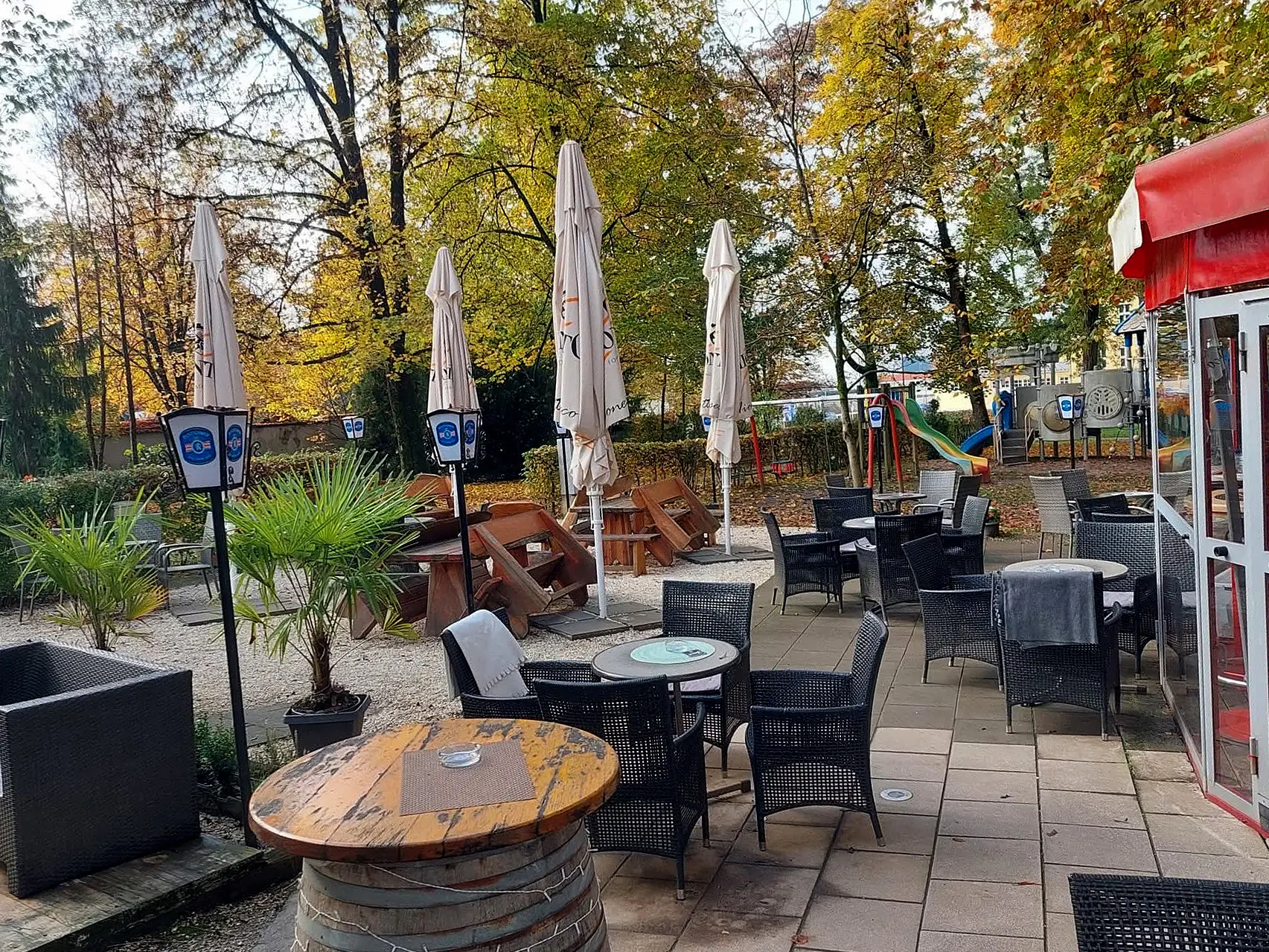 Caféterrasse mit Tischen, Stühlen, geschlossenen Sonnenschirmen und herbstlichen Bäumen