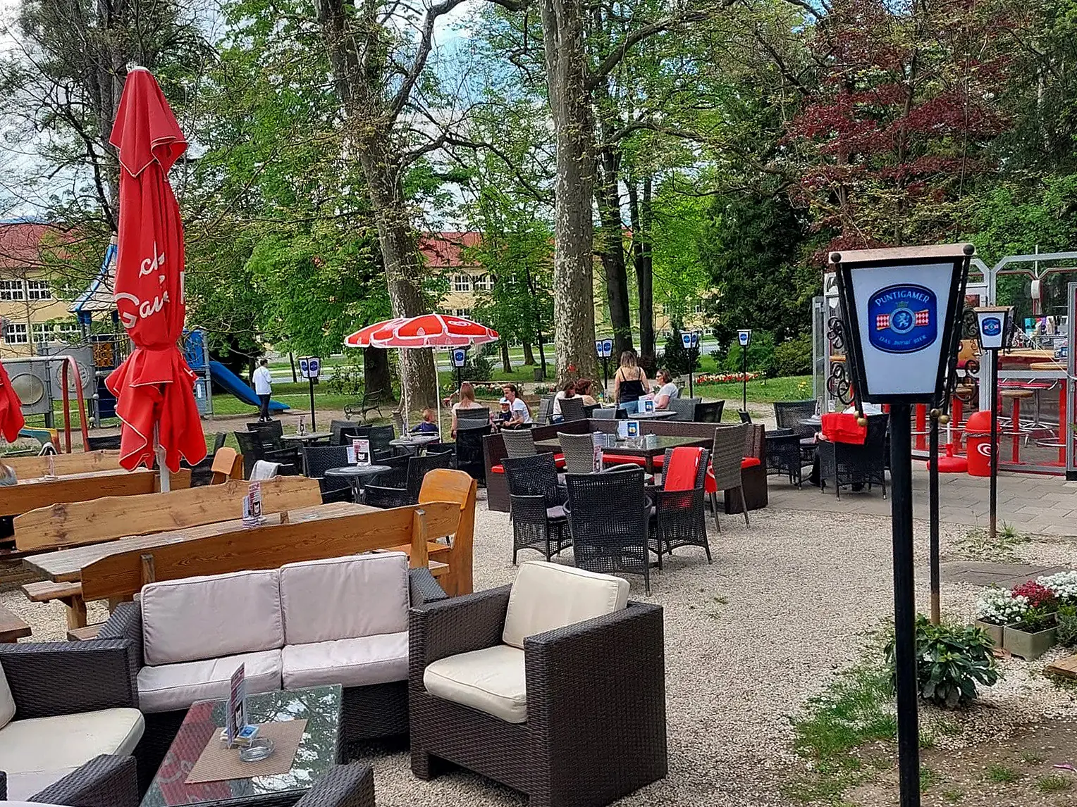 Caféterrasse mit Tischen, Korbsesseln, roten Sonnenschirmen und Gästen unter Bäumen
