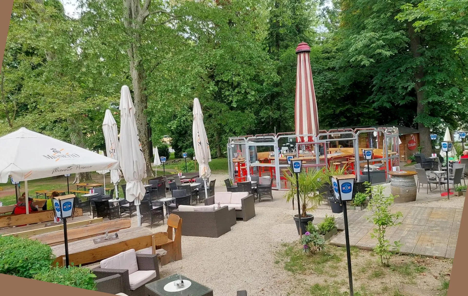 Gartenlokal mit Außentischen, geschlossenen Sonnenschirmen und Sitzgruppen unter hohen Bäumen