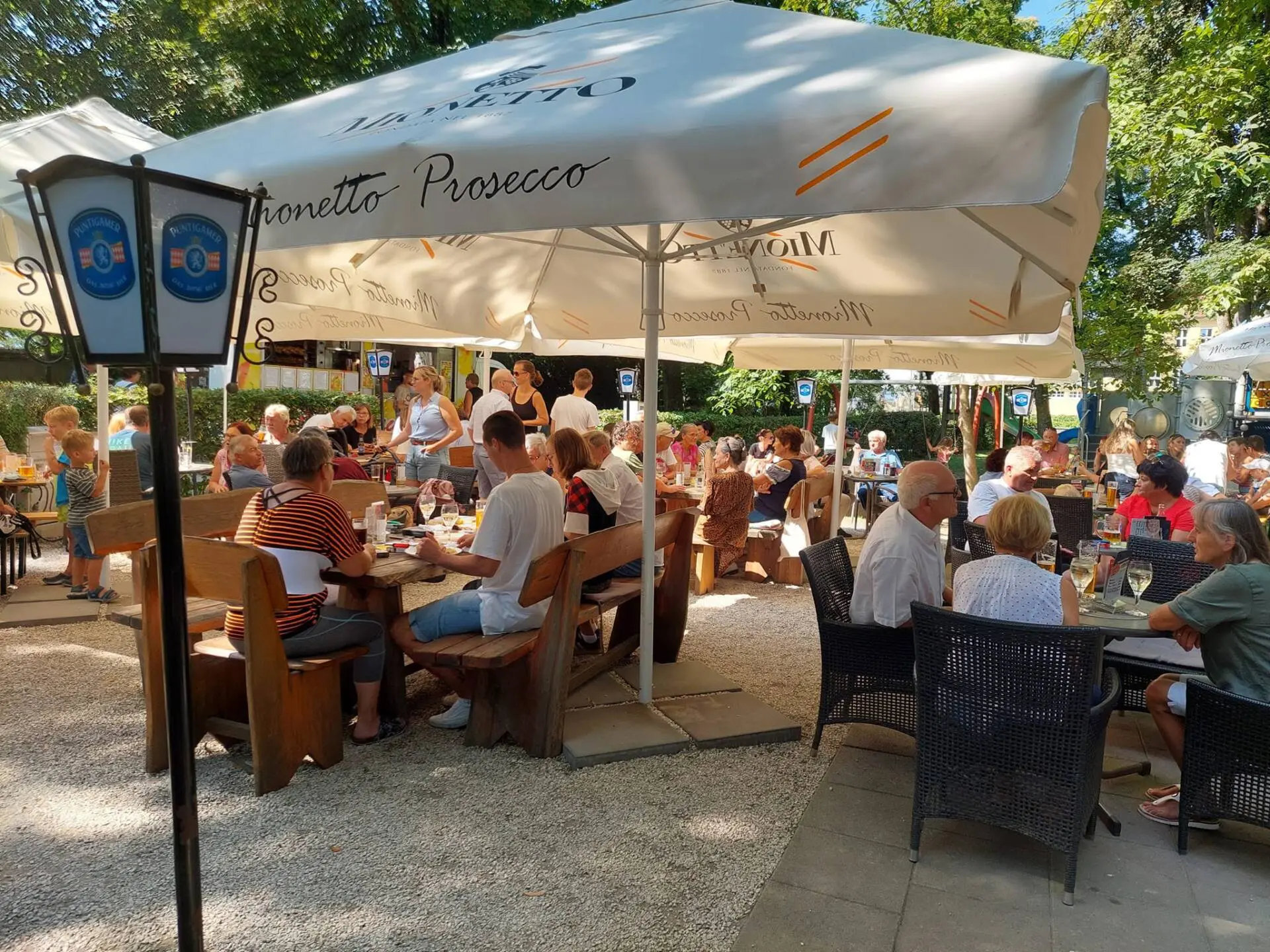 Gäste sitzen an Tischen auf einer schattigen Caféterrasse unter großen Sonnenschirmen.