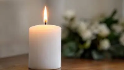 KI-generiertes Bild: Eine brennende weiße Kerze steht vor unscharfen Blumen.