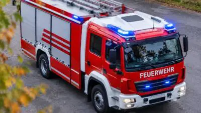 KI-generiertes Bild: Ein rotes Feuerwehrfahrzeug mit Leitern und eingeschaltetem Blaulicht steht auf einer Straße.