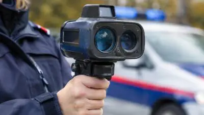 KI-Symbolbild: Eine uniformierte Person hält ein Messgerät vor einem unscharfen Auto mit blauem Lichtbalken.