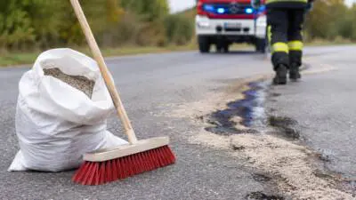 KI-generiertes Bild: Ein Sack und ein Besen liegen neben einer dunklen Spur auf der Straße; im Hintergrund sind eine Einsatzkraft und ein rotes Einsatzfahrzeug zu sehen.