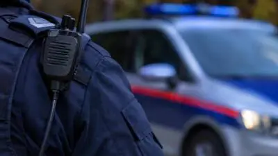 KI-Symbolbild: Eine dunkel gekleidete Person trägt ein Funkgerät; dahinter ist ein blaues Fahrzeug mit Lichtbalken und rotem Streifen zu sehen.