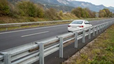 KI-generiertes Bild: Ein weißer Pkw steht nahe einer Leitplanke auf einer Autobahn vor Bergkulisse.