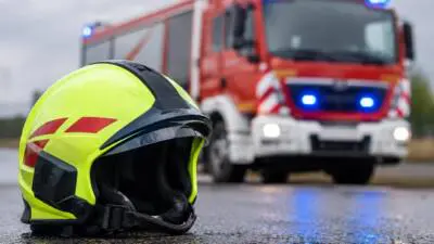 KI-generiertes Bild: Ein gelber Schutzhelm liegt auf nasser Fahrbahn vor einem roten Einsatzfahrzeug mit Blaulicht.