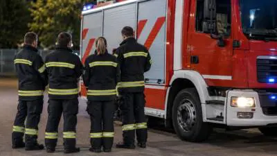 Personen in dunkler Schutzkleidung mit Reflexstreifen stehen neben einem roten Löschfahrzeug.