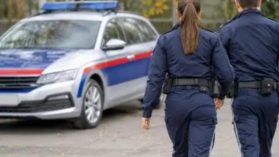 KI-Symbolbild: Uniformierte Personen gehen an einem weiß-blauen Auto mit Blaulicht vorbei.