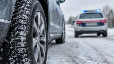 KI-generiertes Bild: Ein Auto mit Winterreifen auf verschneiter Straße, dahinter ein unscharfes Fahrzeug mit blauem Licht.