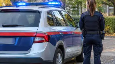 KI-Symbolbild: Eine uniformierte Person steht neben einem blau-weißen Einsatzwagen mit eingeschaltetem Blaulicht.