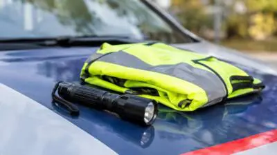 KI-generiertes Bild: Eine Warnweste und eine Taschenlampe liegen auf der Motorhaube eines blau-weißen Fahrzeugs.