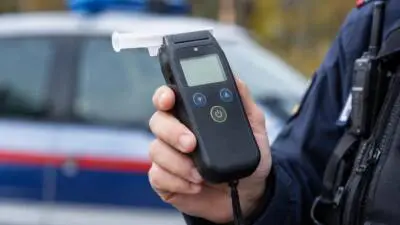 KI-generiertes Bild: Eine Hand hält ein Atemalkoholmessgerät vor einem unscharfen Auto.