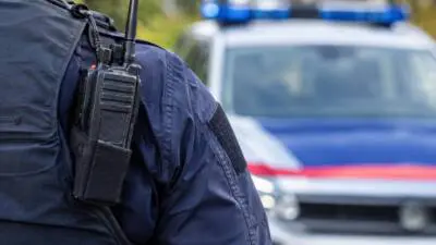 Eine dunkelblau gekleidete Person trägt ein Funkgerät vor einem unscharfen blau-weißen Fahrzeug.