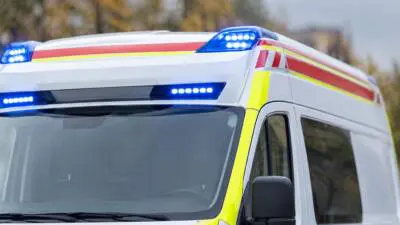 KI-generiertes Bild: Ein weißer Rettungswagen mit eingeschaltetem Blaulicht vor Bäumen.