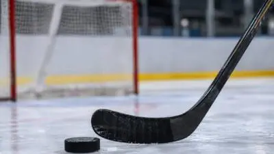 KI-generiertes Bild: Ein schwarzer Eishockeyschläger liegt neben einem Puck auf dem Eis, dahinter steht ein rotes Tor.