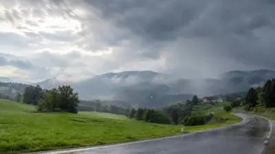 KI-Symbolbild: Eine nasse Landstraße führt durch grüne Hügel unter dunklen Wolken und Regen.